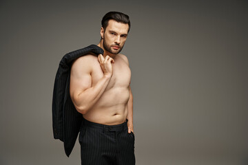 man in his 30s posing with bare chest in pinstripe suit and holding blazer on grey, hand in pocket