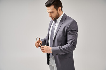 handsome businessman in formal wear holding glasses and standing on grey background, elegance
