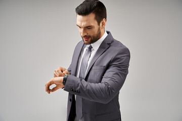 handsome businessman posing in elegant suit checking his wristwatch on grey background, punctual