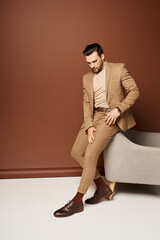 elegant and good looking man in stylish suit standing near armchair on beige backdrop, smart casual