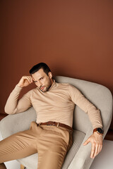 pensive man in turtleneck sitting on comfortable armchair and looking at camera on beige backdrop