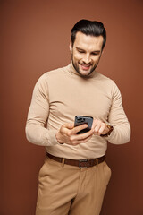 happy man with bristle in turtleneck using his smartphone on beige background, social media