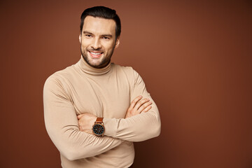 happy and handsome man in turtleneck posing with crossed arms and sharp gaze on beige backdrop