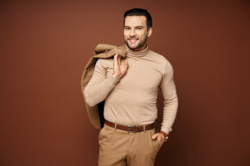 happy man in turtleneck holding his jacket over shoulder and posing with hand in pocket on beige