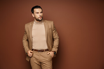 handsome man in elegant attire looking away while posing with hand in pocket on beige backdrop