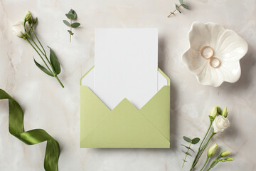 Olive envelope with blank paper card inside, gold rings, green ribbon and flowers on stone...