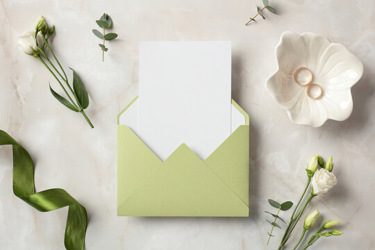 Olive envelope with blank paper card inside, gold rings, green ribbon and flowers on stone background. Wedding invitation card template. Flat lay, top view
