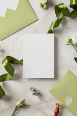 Blank wedding invitations card mockup with space for text, green ribbon, olive envelopes, flowers on stone background.