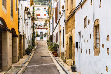 A narrow street with traditional white houses adorned with potted plants, in the old town of the picturesque coastal town of Tossa de Mar, Costa Brava, Spain.