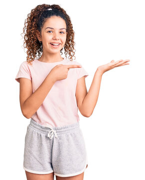 Beautiful kid girl with curly hair wearing casual clothes amazed and smiling to the camera while presenting with hand and pointing with finger.