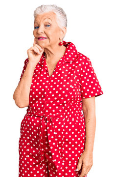 Senior beautiful woman with blue eyes and grey hair wearing a red summer dress with hand on chin thinking about question, pensive expression. smiling with thoughtful face. doubt concept.