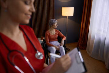 Doctor caregiver making notes about old senior woman patient - obrazy, fototapety, plakaty