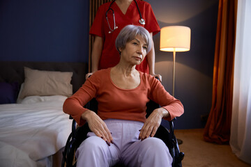 Aged senior woman in wheelchair having professional help at nursing home - obrazy, fototapety, plakaty