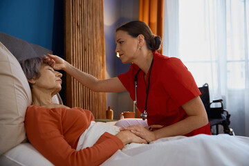 Female caregiver measuring temperature touching forehead of old woman - obrazy, fototapety, plakaty