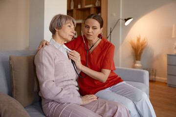 Woman doctor using stethoscope while examining senior woman at nursing home - obrazy, fototapety, plakaty
