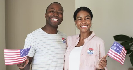 A diverse couple is beaming with pride, holding American flags and wearing vote stickers