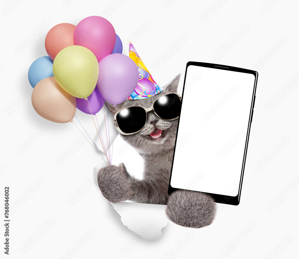 Poster happy kitten wearing party cap holds bunch of balloons, blows in party horn and shows smartphone wit