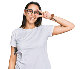 Young hispanic woman wearing casual white t shirt doing peace symbol with fingers over face, smiling cheerful showing victory