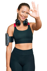 Beautiful blonde woman wearing gym clothes and using headphones smiling positive doing ok sign with...