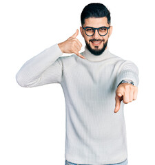 Fototapeta premium Young arab man with beard wearing elegant turtleneck sweater and glasses smiling doing talking on the telephone gesture and pointing to you. call me.