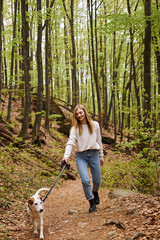 Naklejka na ściany i meble Laughing joyful blonde hiker girl walking dog in woods in forest while trekking, adventure