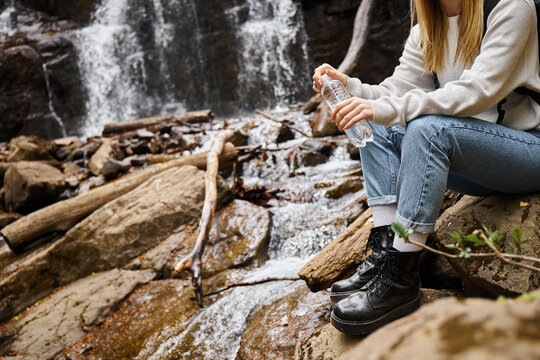 Cropped image of woman in jeans sitting with bottle of water on rocks in forest near waterfall