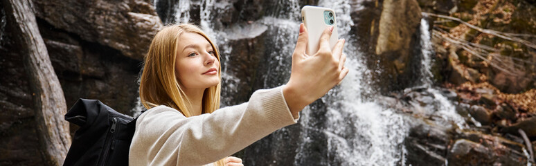 Blonde young woman taking selfie near mountain waterfall in the forest while hiking, banner