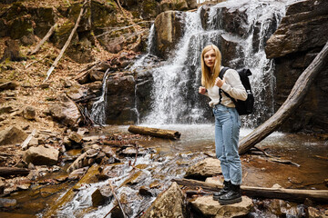 Active blonde female hiker standing by the forest creek walking on rocks near waterfall
