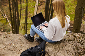 Back view of blonde female hiker sitting on boulder in forest with laptop on her knees with backpack