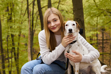 Smiling blonde woman hugging her dog, while having a halt on forest trip, looking at camera