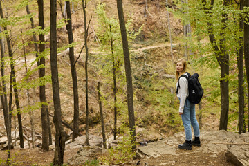 Side view of blonde young woman adventurer standing on forest trail, discovering new paths