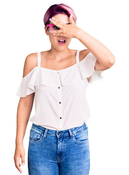 Young beautiful woman with pink hair wearing casual clothes and glasses peeking in shock covering face and eyes with hand, looking through fingers with embarrassed expression.