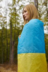 Side view of blonde young woman wrapping the Ukrainian flag over her body in forest scenery