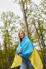 Patriotic blonde girl proudly holding national Ukrainian flag in forest and looking at camera