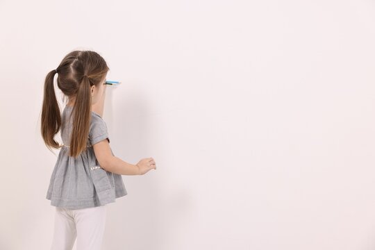 Little girl drawing with colorful pencils on white wall indoors. Space for text