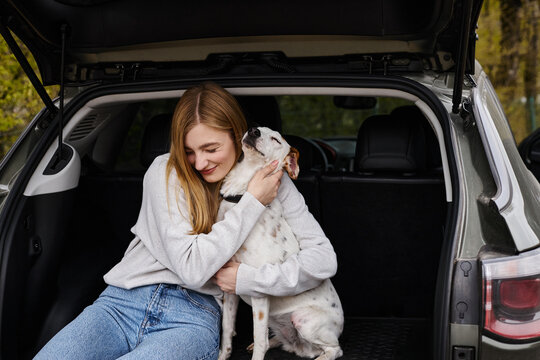 Image of woman gently hugging her white dog sitting at back of car. Dog companion