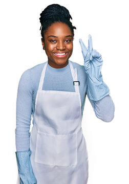Young african american woman wearing apron smiling with happy face winking at the camera doing victory sign. number two.