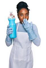 Young african american woman wearing apron holding detergent bottle covering mouth with hand, shocked and afraid for mistake. surprised expression