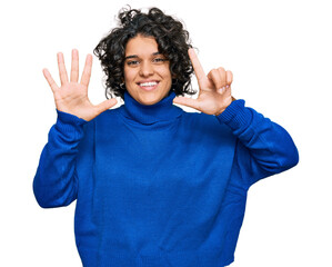 Young hispanic woman with curly hair wearing turtleneck sweater showing and pointing up with fingers number seven while smiling confident and happy.