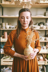 Creative Woman Potter with Clay Vase in Artisan Studio with eyes closed. Artistic woman enjoying her hobby, holding a unique piece of pottery in a well-lit studio