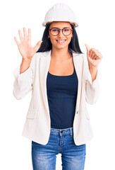 Obraz na płótnie Canvas Young beautiful latin girl wearing architect hardhat and glasses showing and pointing up with fingers number six while smiling confident and happy.