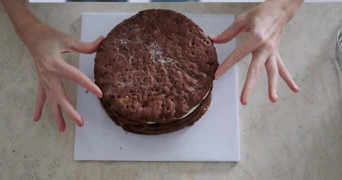 Close-up shot from top to bottom on the hands of a young female pastry chef applying raspberry jam to a chocolate cake. The process of preparing layers of cake in the kitchen in a pastry shop.