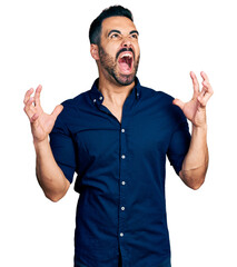 Young hispanic man with beard wearing casual blue shirt crazy and mad shouting and yelling with aggressive expression and arms raised. frustration concept.
