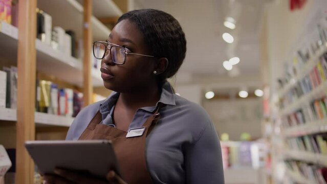 Medium footage of young African American female makeup store employee in glasses and apron standing on shop floor, counting stocks of merchandise on shelves, and ordering new supplies