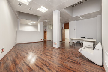 An empty office with plain white painted walls, a pillar in the middle, skylights on the ceiling, dark wood door carpentry and skirting boards, reddish parquet floors and technical ceilings