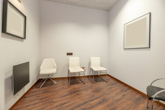 An empty office with plain white painted walls, white skay upholstered seats, reddish parquet floors and technical ceilings