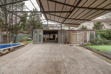Fototapeta premium A covered terrace on the plot of a single-family home with a canvas roof on a metal structure and a room with aluminum doors and a vehicle parked inside.