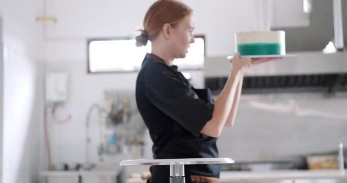 A young charming red haired female pastry chef takes a cake from the table in the industrial kitchen of a pastry shop and takes it to the refrigerator. The process of preparing sweet desserts.