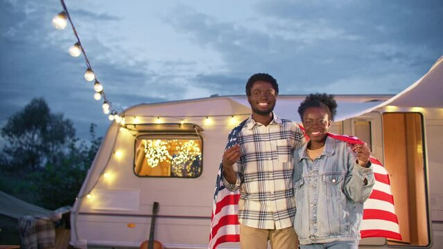 Zooming In Positive African American Couple Covered With Flag Of United States. People Hugging Each Other While Joyfully Smiling At Camera. Behind People Located Camper Van. Light Bulbs Glowing Around