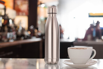 One metal vacuum-insulated bottle for water or coffee and a cup of coffee on a table on a bar background. - obrazy, fototapety, plakaty